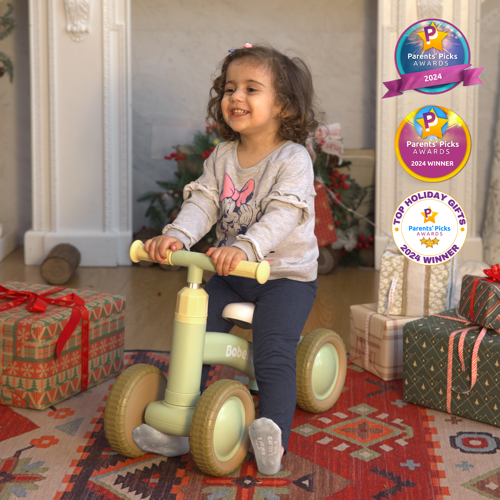 Smiling toddler riding a Babelio balance bike in a living room with gifts and holiday decor