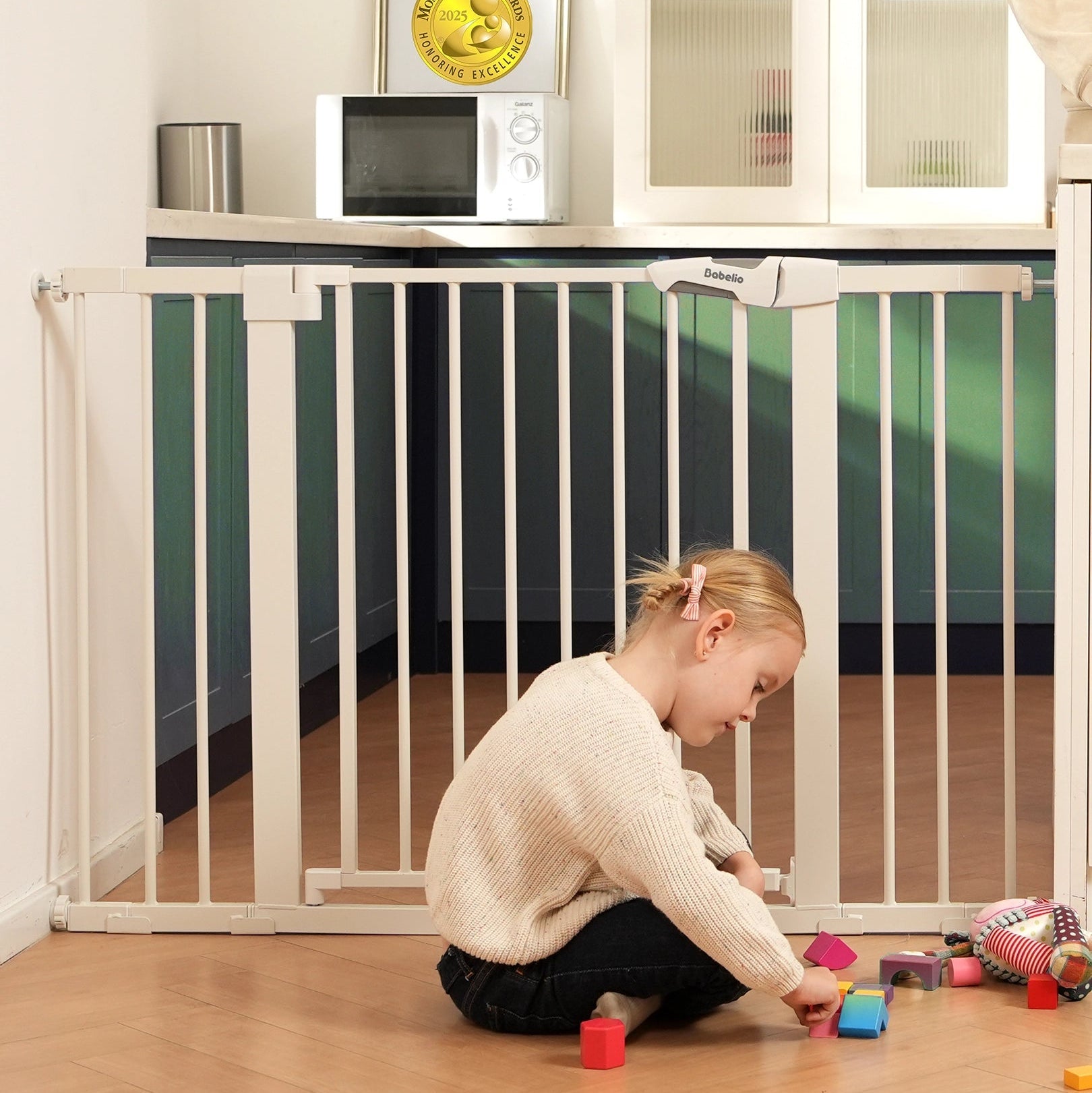 Babelio baby safety gate installed in a kitchen, with a child playing on the floor nearby.#Color_White
